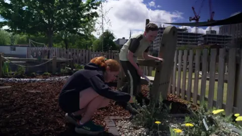 Two children are tending to plants in a rain garden. A boy is standing next to a wooden chair while the a girl crouches next to a small shrub.