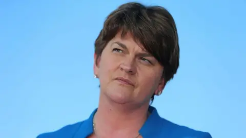 Arlene Foster stares upward and to the right. She has short, dark hair parted to the side and is wearing a blue jacket