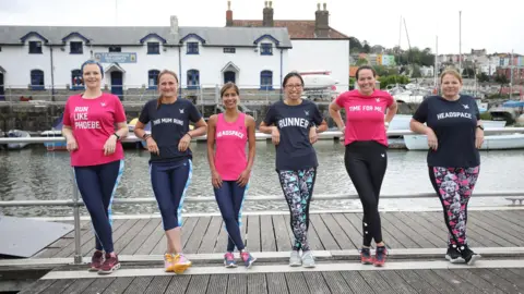 This Woman Runs A group of six women stand on Bristol's harbourside, with some of the city's rows of iconic colourful houses in the background, and they smile at the camera. They all wear running gear and trainers.