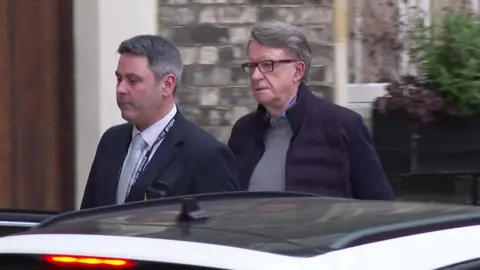 BBC Lord Mandelson is led towards a car by a police officer who is not in uniform
