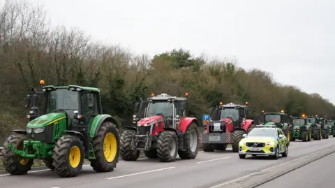 Shaun Whitmore/BBC Tractors are pictured moving along a dual carriageway in a single line formation while a police car drives along side them in the parallel lane. 