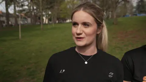 A woman with blonde hair tied in a ponytail in a park with green grass behind her. She is wearing a black top and silver necklace