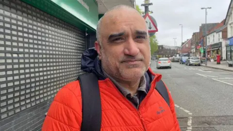 A man stands in a high street in front of a shop with its shutters down. He has a bright red jacket and is wearing a backpack with black straps. He has closely shaved grey and black hair and stubble.