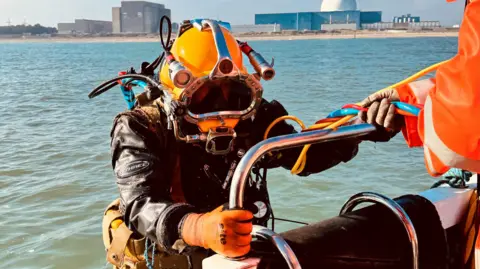 EDF Energy A diver holding onto the side of a platoon floating in the sea. The diver is wearing a diving suit and has there is someone in a high-visibility orange jacket helping him. 