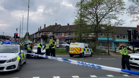 BBC/Cathy Killick A police cordon cuts across Otley Road in Headingley. Police officer in high visibility jackets are stationed on the road. Three police vehicles are parked on the road. A tree stands in front of a line of shop fronts behind.