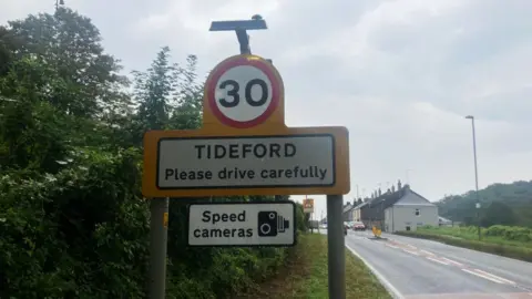 BBC A road speed limit sign which says "30", and, below it 'Tideford, please drive carefully". Below that is a sign saying "speed cameras" and a black and white graphic of a camera. Beside the sign on the right is the road.