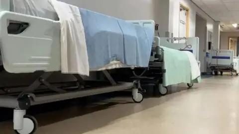 Empty hospital beds lining the corridor of a hospital. They are on wheels and have blue and green blankets draped over them.