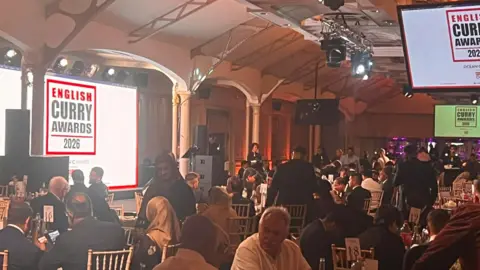 Basmati Restaurant A room featuring dozens of people sitting down and large mainly white screen on the left including the words English Curry Awards 2026.