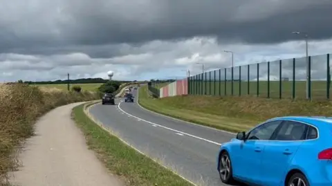Several cars, including a blue hatchback, drive along a road on the boundary of a military base in Cornwall. The base has large fencing - including several sections coloured red and white - marking out the border. It is an overcast day.