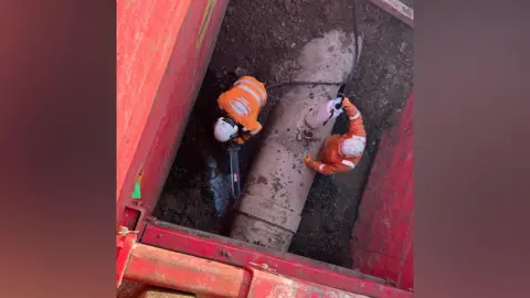 Northumbrian Water An overhead view of two workers working on a large, brown sewer pipe. They are wearing orange high-vis jackets and white helmets. The pipe is surrounded by mud. One worker appears to be holding a chainsaw.