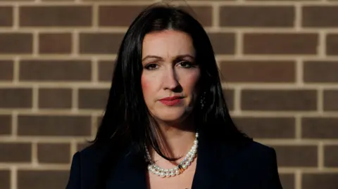 PA Media A head shot of Emma Little-Pengelly, a woman with black hair wearing a pearl necklace and a black top, is standing in front of a brick wall.