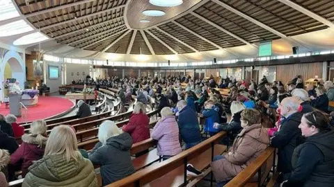 Rory Healy A church hall packed with mourners - it is a modern design of church with the pews in a semi-circular shape. 