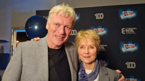 David Moyes and Jenny Seagrove pose together in front of a board with branding of the theatre and Blood Brothers in the lobby