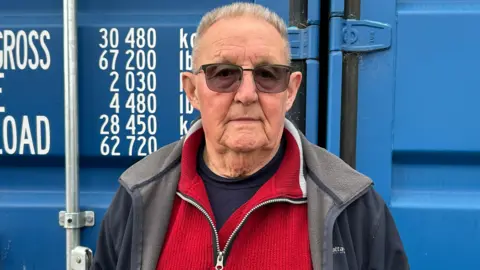 An elderly man with short grey hair and sunglasses, wearing a red sweater under a dark blue and grey fleece. He is standing in front of a blue shipping-style container.