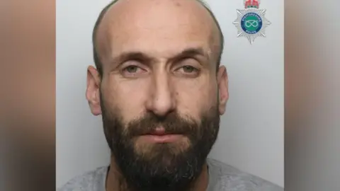 A man with short hair and a beard, in front of a white wall. The Staffordshire Police badge is in the top right corner.