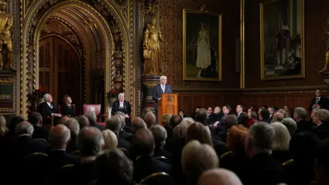 Reuters Steinmeier stands at a wooden lectern in an ornate hall as rows of seated men and women watch on.