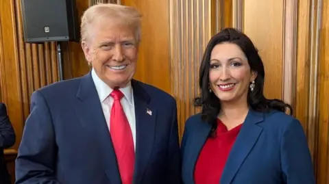 NI Executive Donald Trump poses with Emma Little Pengelly. He wears a navy suit over a white shirt and a red tie. She wears a blue jacket over a red dress. They stand in a wooden paneled room. Trump has blonde/white hair, while small Pengely has long dark hair.