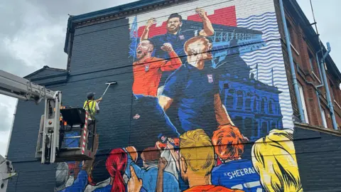 Alice Cunningham/BBC A painter on a platform lift paints over a mural that depicted Ipswich Town's players and manager on the side of a building.