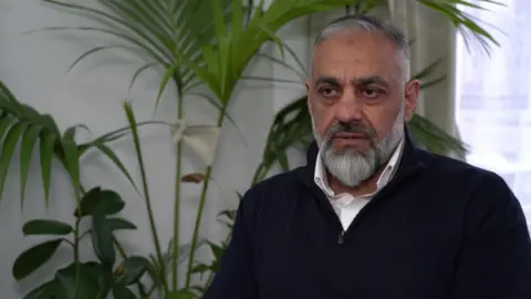 Shahid Butt, who has grey hair and a grey pointed beard, is wearing a dark zip up top and a white shirt underneath and is sitting down being interviewed. There is a white wall with a green plant behind him, and a window with a beige curtain. 