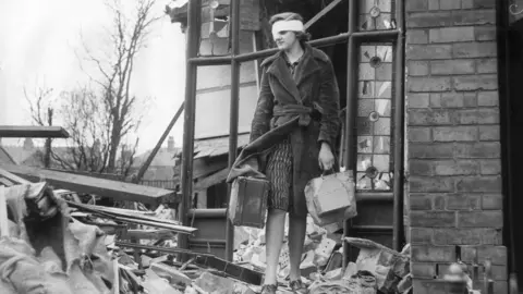 A black-and-white photo showing a woman with a bandaged head carrying a suitcase and a bag as she clambers over rubble outside her bomb-damaged home. The door is broken, widows smashed and wooden frames askew.