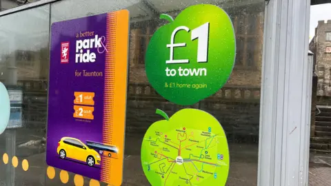 Three large stickers on a bus stop's glass advertising Taunton's Park and Ride services. Two of the stickers are shaped like apples and one of them says "£1 to town & £1 home again" on it. The other is a purple and orange rectangular sticker that says "a better park and ride for Taunton" with photos of tickets and a yellow car.