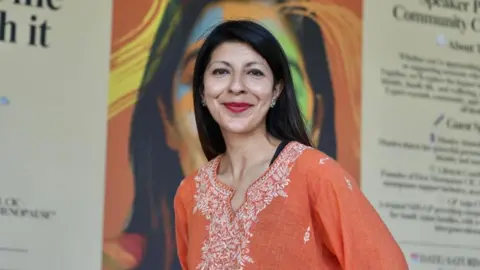 Colin Moody Sheetal Jethwa, founder of South Asian Voices Bristol CIC, poses for a picture in front of large posters. She is wearing an orange top with white embroidery around the neckline and red lipstick. She has long, dark hair.