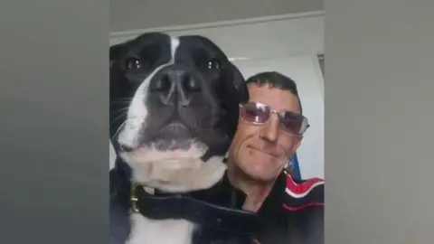 Suffolk Police A dark-haired man wearing glasses and a black and red top, sitting with a black and white dog.