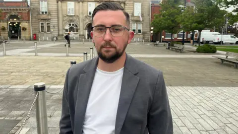 A man with short brown hair, glasses, a beard, a grey jacket and white shirt, stands in the middle of a town square. There is a large, brown stone building in the background. 