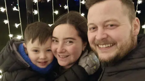 Supplied The family of Maxwell, Viki, and Luke are smiling with what looks to be Christmas lights in the background. 