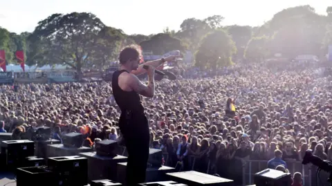 Tom Grennan performs at Radio 1's Big Weekend on Saturday 27 May 2023. He is standing on stage singing into a microphone in front of a vast sea of people below him.