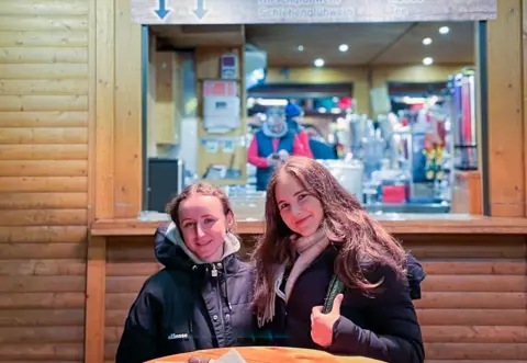 Two young women sit at a wooden table in front of a wooden hut selling chocolate covered strawberries at a Christmas market in Berlin. They are both smiling.