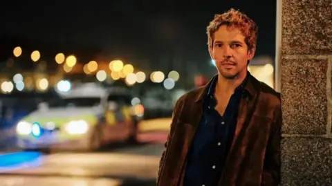 Escena nocturna callejera de UKTV en la que el actor Damien Molony aparece apoyado en la pared de un edificio de piedra. Está enfocado, vistiendo una camisa oscura y una chaqueta marrón, mientras que un vehículo policial con las luces encendidas se ve borroso al fondo. Las cálidas luces de la ciudad y los reflejos crean una atmósfera cinematográfica con poca profundidad de campo.