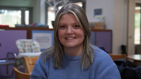 John Fairhall/BBC Hannah Taylor, is a woman who is sitting in an EACH hospice. She is looking at the camera and is smiling. She has blonde hair and is wearing a light blue jumper.