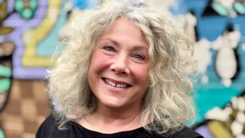 Gemma Dillon/BBC Image shows a lady wearing a black top with wavy grey hair.
She is standing in front of a wall with modern art grafitti.