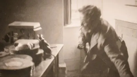 Jeff Towns Dylans Bookstore Dylan Thomas in a black and white photo standing beside the same window but is crouching with the chair legs still in one hand. A table beside him has audio recording equipment from the era and film cans stacked up.