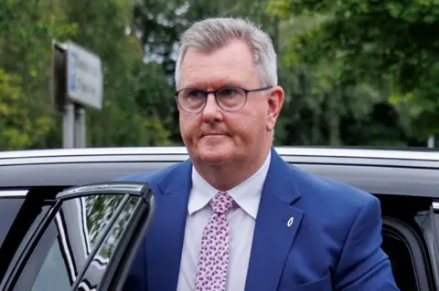 Jeffrey Donaldson is walking away from a black car. He is a man with short grey hair, wearing a dark blue suit, white shirt and pink tie.