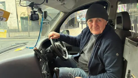 Thomas Berrington/BBC Arthur Grimes, wearing a dark grey beaie hat, blue zip-up fleece and grey jumper, smiles at the camera from the driver's seat of a black cab. His right hand is on the steering wheel. 