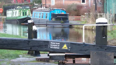 John Sadler Blue and green narrowboats are on the canal at Stone near a lock. The picture shows the black and white lock close up with a sign that says "keep boat forward of cill marker". There are buildings by the canal.