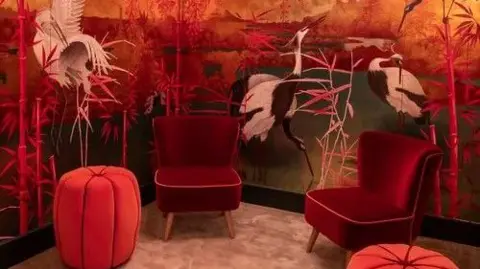 The Sherborne Tropical birds and plants on the walls of the washroom with red and yellow tropical scenes across the walls. There are red velvet chairs on the ground.