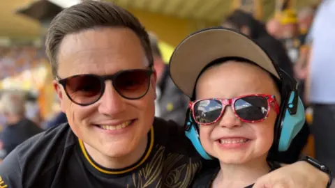 Kieran Newey Kieran Newey is pictured with his son at a football match. They both have sunglasses on and are beaming at the camera. His son has blue ear defenders on and a cap. Kieran has a Wolves top on.