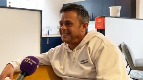 A man is smiling sitting on a yellow sofa. He is wearing a white hoodie with a black adidas logo. There is a purple microphone being held in front of him reading BBC RADIO STOKE. An open-plan kitchen and a chair can be seen in the background.