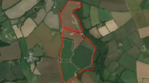 An aerial/birds eye view shot of the proposed solar farm site. The area is rural with lots of green open fields and trees. The site appears to cover six fields and is marked out clear in red. 