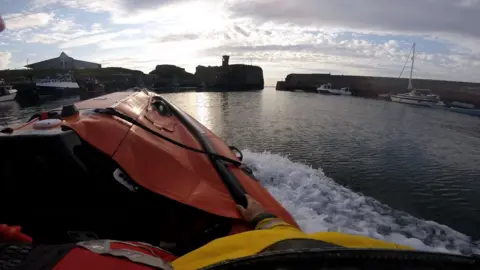 Photo taken from a lifeboat showing the boat heading out of a harbour where there are other boats moored.