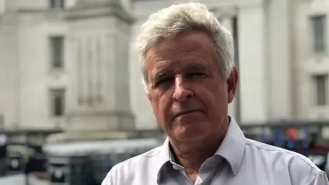 Steve Goodman with short white hair looking at the camera and wearing a pink shirt. He is standing in front of shops in Luton.