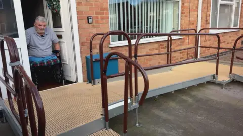 Neville Bradford sitting in his wheelchair in his house doorway looking at the ramp outside his home which slopes down to the right underneath his window. It has burgundy railings and a separate step in the foreground.