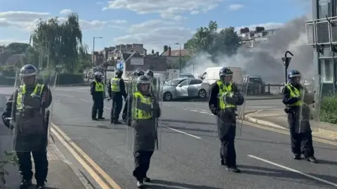 A group of riot police wearing helmets and hi-vis vests hold clear plastic shields as they guard a street. In the background, plumes of smoke rise from a car park and a damaged car can be seen.