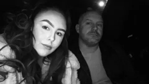 A black and white picture of Mollie Lord (left) and her dad Warren. She has long dark hair and wears a light coloured faux fur-style jacket. He has a bald head and wears a t-shirt and a black jacket