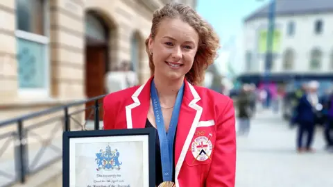 Hannah Scott is standing outside Coleraine Town Hall.  She is wearing a red blazer with a white trim around the lapels and cuffs. She has blond curly hair and is holding a framed certificate of her freedom of the borough