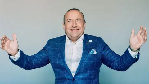 Dave Fishwick stands smiling widely with his arms outstretched to the sides/ He has very short dark hair and is wearing a blue checked suit jacket and a white shirt