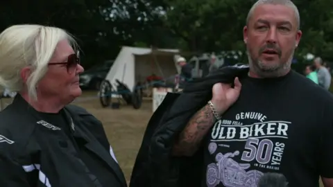 A man and woman wearing black biker jackets over a black T-shirts. The woman is wearing sunglasses and has blonde hair, while the man has grey shaved hair and a short dark beard with hints of grey.